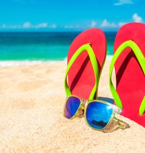 Flip floops and sunglasses on a beach with the sea in the background representing summer holidays