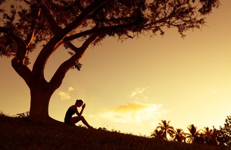 Woman sitting under tree worrying - shadowed image with sunset background