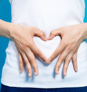 Torso of a woman in white t-shirt making the shape of a heart with her hands, over her stomach.