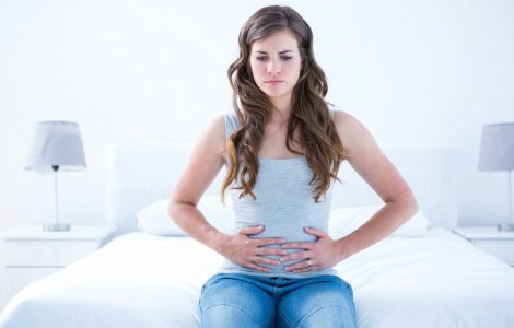 Young woman sat on a bed clutching her stomach to represent tummy ache