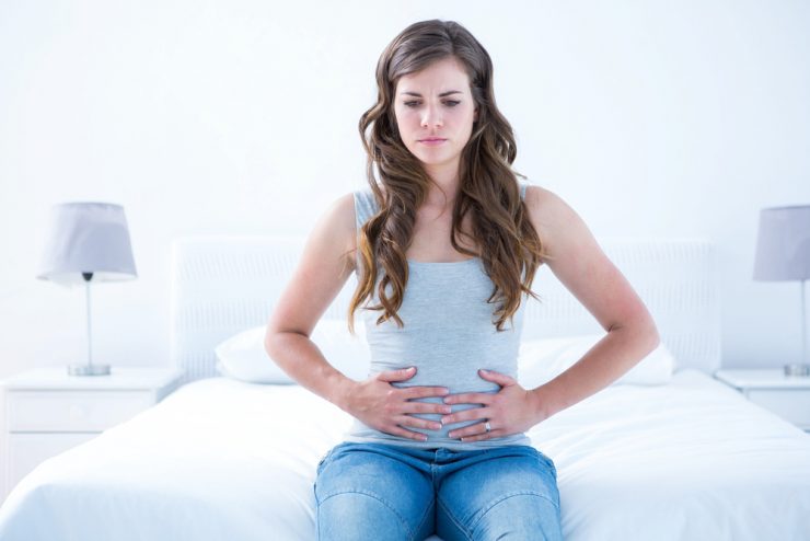 Young woman sat on a bed clutching her stomach to represent tummy ache