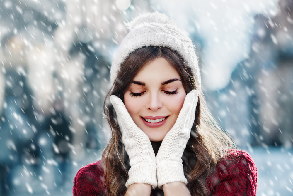 Close up of woman outdoors in the snow with her hands in gloves by her face to show winter skincare